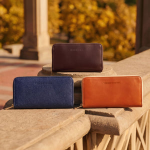 Three leather zip-around wallets are arranged on a stone railing outdoors with a blurred autumn park background.