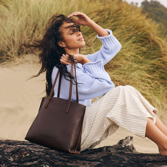 A model sitting on a log on the beach holding a Coldbrew August Tote.