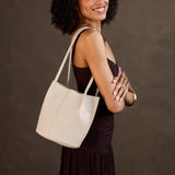 Bone Small | Model wearing sleeveless dark brown dress carrying petite bucket shaped tote bag with matching leather handles against dark grey background