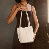 Bone Small | Model holding sleeveless dark brown dress carrying petite bucket shaped tote bag with matching leather handles against dark grey background