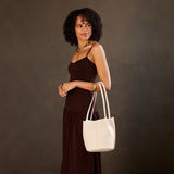 Bone Small | Model wearing sleeveless dark brown dress carrying petite bucket shaped tote bag with matching leather handles against dark grey background