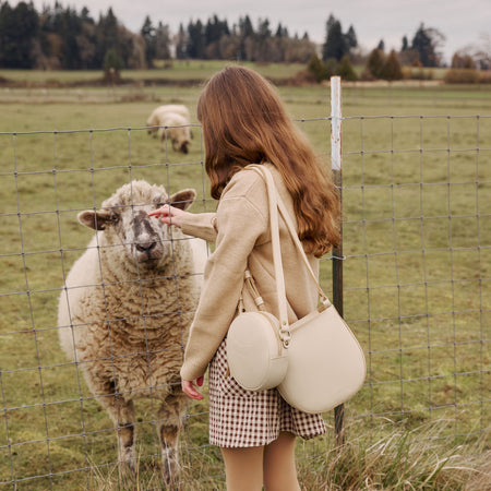 Child wearing a Petal Crossbody and Sunnie Crossbody in Pebbled Bone while petting a sheep through a fence