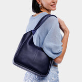 Cobalt Large | A model holding a u-shaped, flat-bottomed leather shoulder bag with two outside pockets
