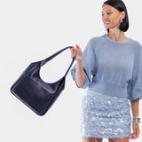 Cobalt Large | A model holding a u-shaped, flat-bottomed leather shoulder bag with two outside pockets