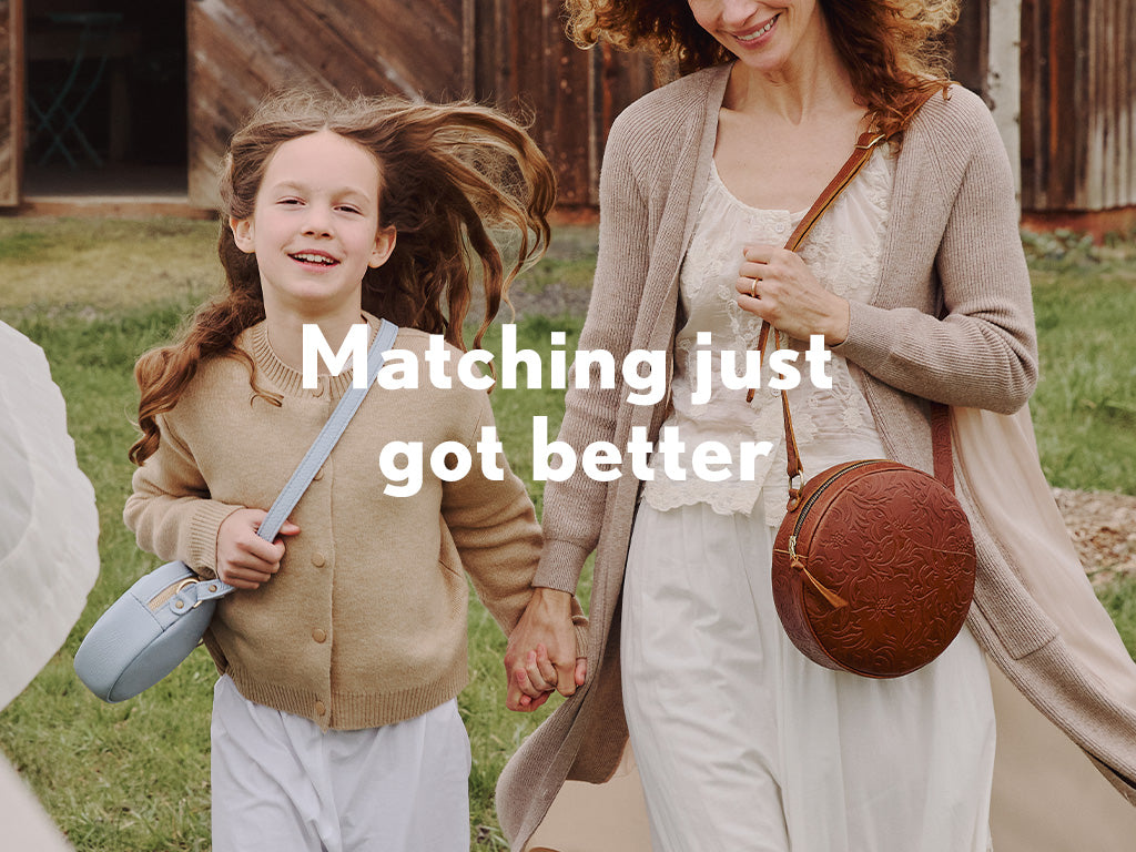 Woman and child holding hands with coordinating outfits. Child is wearing a Sunnie Crossbody in Skyway, woman wearing a Circle Crossbody in Meadow. Text says matching just got better
