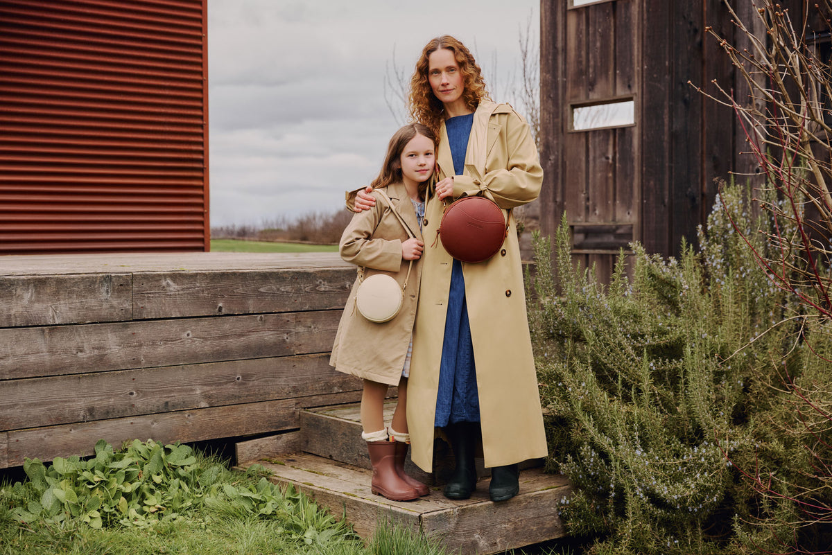 A women and a child standing on wooden steps outdoors, wearing beige coats and wearing a Circle Crossbody bag in Nutmeg and a Sunnie Crossbody in Pebbled Bone