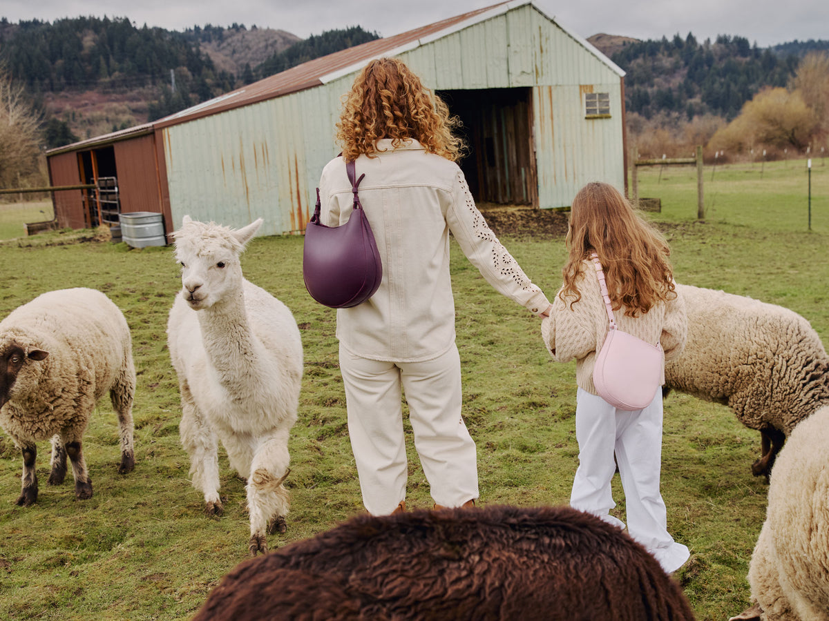 A adult and child in white outfits carrying the Olivia Shoulder bag in Plum and the Petal Crossbody in Pinkaboo standing among sheep in a pastoral setting.