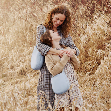 Woman hugging a child in a field of tall grass, both wearing plaid and floral outfits. The woman is holding a Olivia Shoulder bag in Skyway and the child is holding the Petal Crossbody in Skyway.