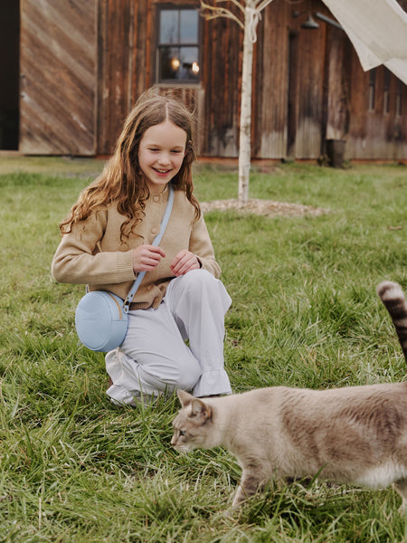 Young child wearing a Sunnie Crossbody in Skyway sitting on grass with a cat in front of a wooden building