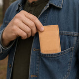 Naturale |A close-up of a model wearing a denim jacket and black shirt, holding a tan leather card holder in their hand. 