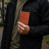 Toscano | A man wearing a black jacket and olive green shirt holds a brown leather card holder in his hand in an outdoor setting.