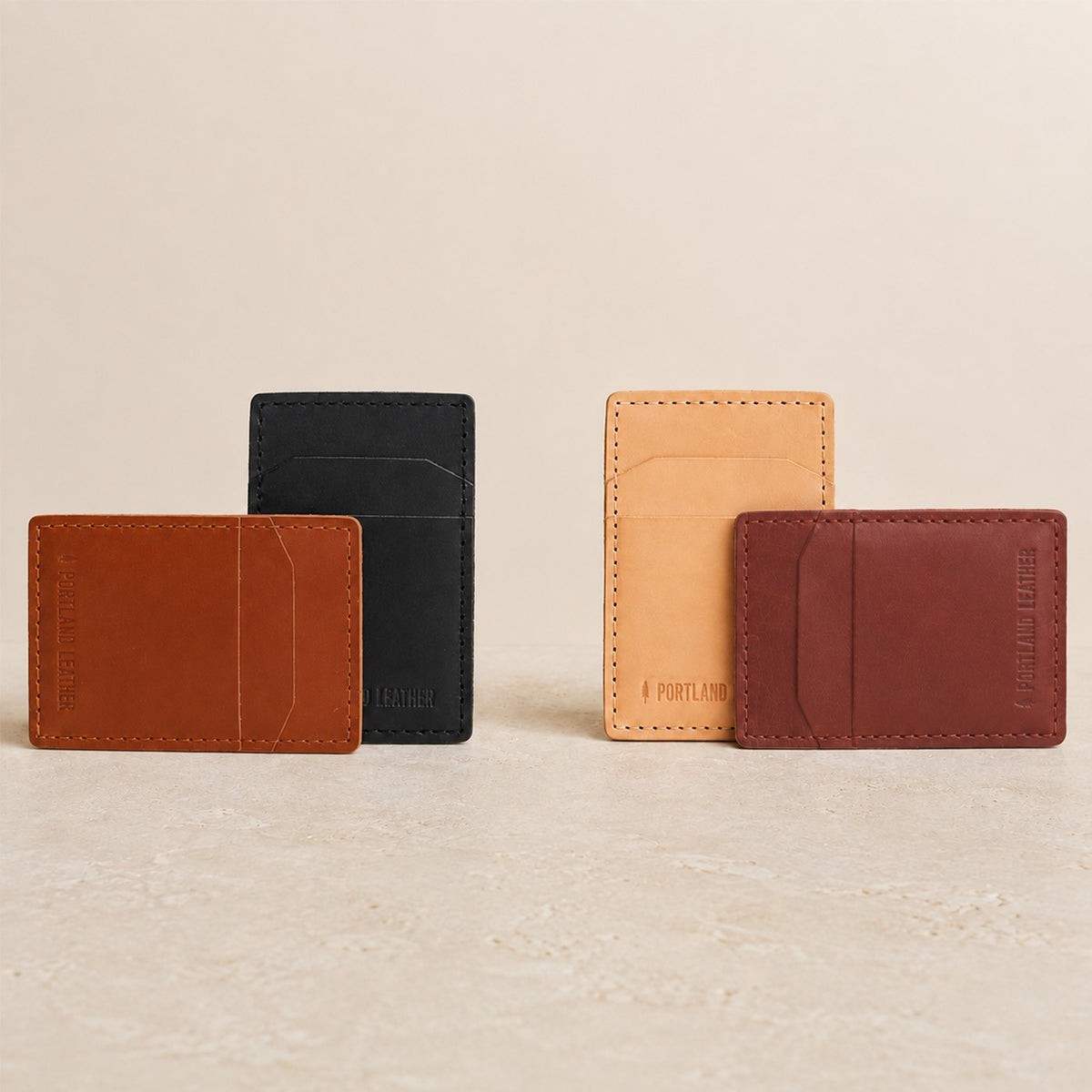 Toscano | Four slim leather card holders in brown, black, tan, and dark brown, arranged in pairs on a light beige surface against a matching neutral background. 