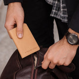 Naturale | A close-up lifestyle shot of a person's hands holding a card holder near a brown leather bag
