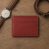 Marrone | A leather card holder displayed on a wooden surface next to a brown leather watch strap and a braided bracelet