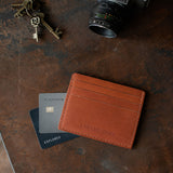 Toscano | A leather card holder styled on a dark textured surface with credit cards, keys, and a camera lens